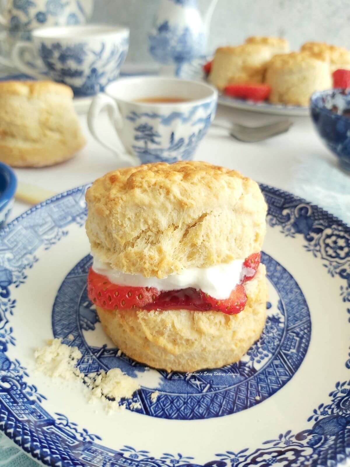 Close photo of a British scone with cream, jam and strawberry sliced in-between, serve don blue & white Willow tea set and more scones and tea to the background.
