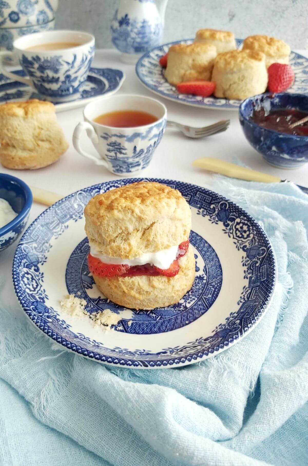 long photo of a British scone with cream, jam and strawberry sliced in-between, served on blue & white Willow tea set.