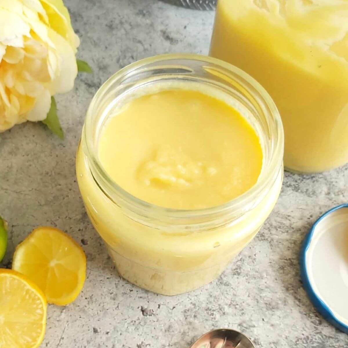 Overhead shot of an open jar of homemade lemon curd. with another jar to the back, lid and lemon slices.