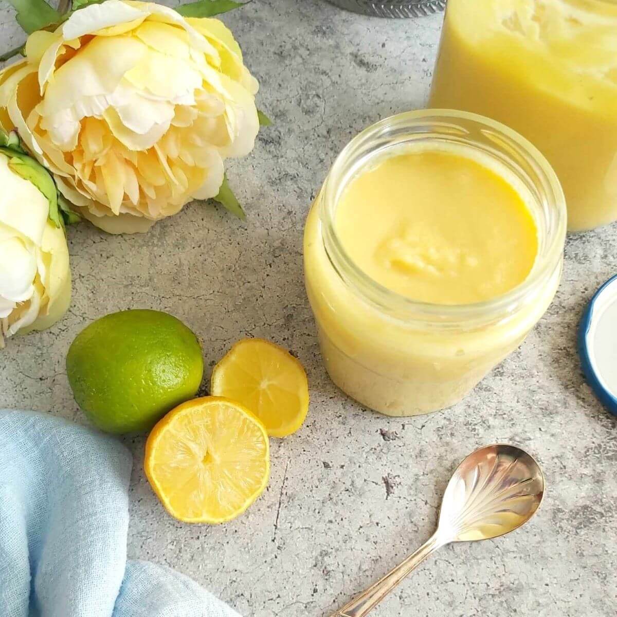 Overhead shot of a open jar of homemad elemon curd, with another jar to the back, lid, lemon and lime slices and yellow coloured flower.