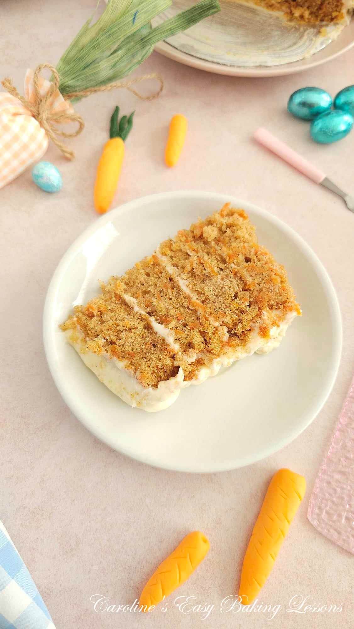 Angled shot of a pale pink table with Easter mini eggs and fondnt carrots and a plate in centre with slice of 4 layer carrot cake with cream cheese frosting.