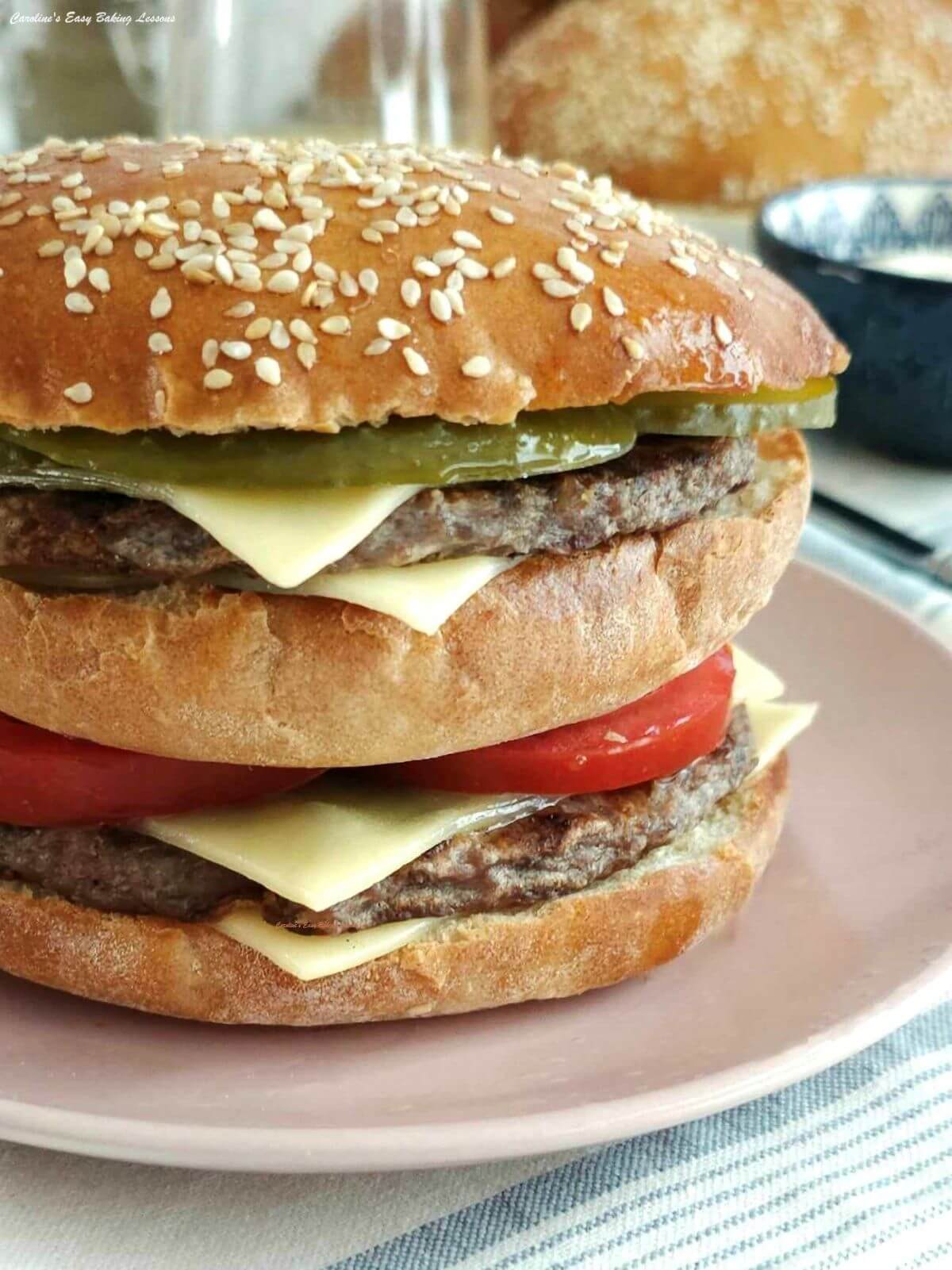 Extra close partial shot of tall homemade double decker cheese burger with deep golden brown seeded top, cheese, tomoato and gerkins visible, on a pale pink plate.