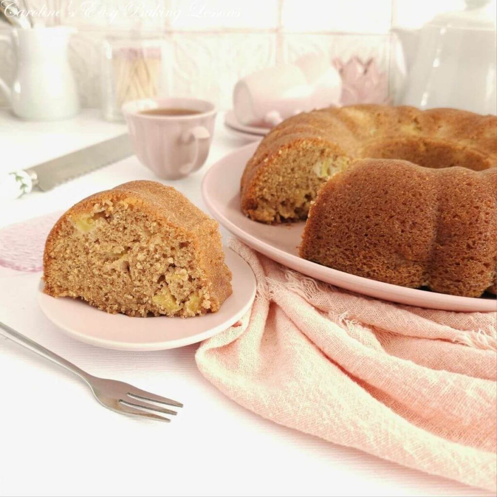 Photo of a slice of apple bundt cake served ona pink plate, with cake to the side, pink napkin and crockery.