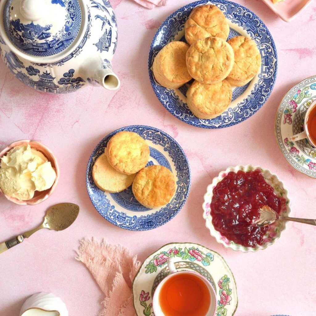 Overhead of a pale pink tabletop with British Cream Tea setting of scones, clotted cream, jam and tea.