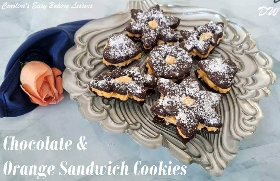 Shot of a decorated silver coloured plate with chocolate sandwich cookies in various shapes, with orange coloured buttercream filling and rose and napkin to the side.