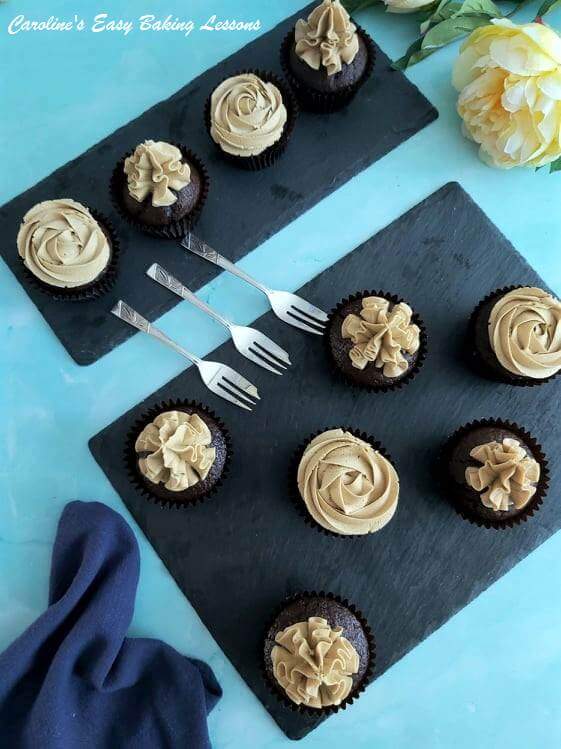 Overhead shot on blue table, and black slate serving trays, with10 chocolate cupcakes, topped with piped espresson buttercream, with forks, npakin and flower.