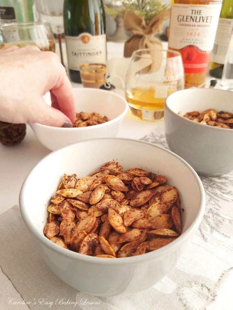 Ladies hand reching into a bowl of nibbles, with drinks and glasses and bowl at front with seasoned pumpkin seeds.