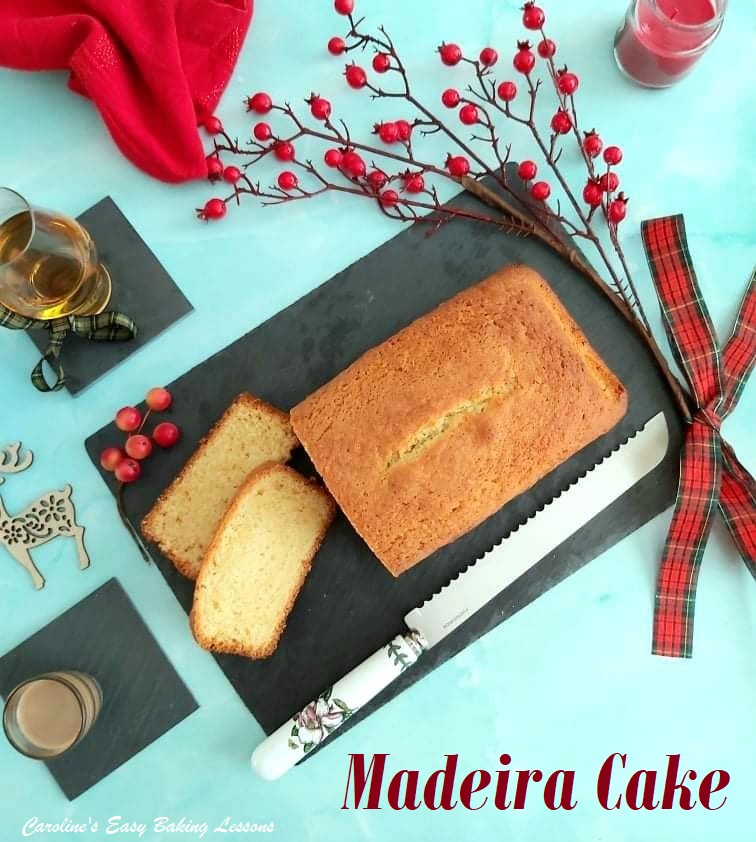 Madeira cake partialy sliced, on top of a black slate board, on a pale blue background, with whisky glass, Baily's drink, fancy knife, red berries, red napkin and Scottish tartin bow.