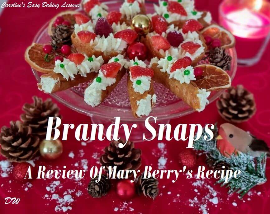front on shot of glass plate of brandy snap tubes wth cream and strawberries pieces with Christmas candle to the side & title brandy snaps Review Of Mary Berry Recipe.