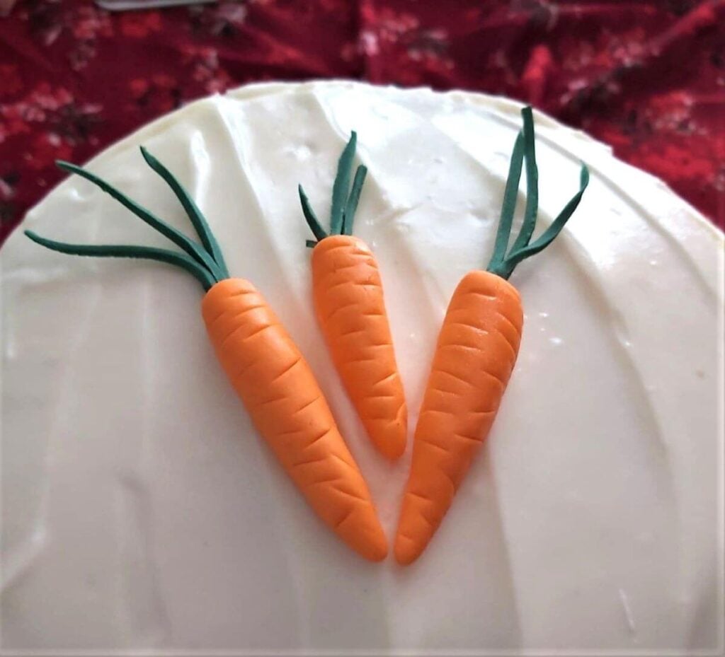 Close shot of handmade fondant carrots on top of asmooth frosted carrot cake.