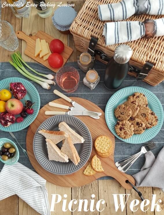 Overhead shot of a picnic table with basket, flask, napkins, sandwiches, cookies, fruit etc and titled picnic week.