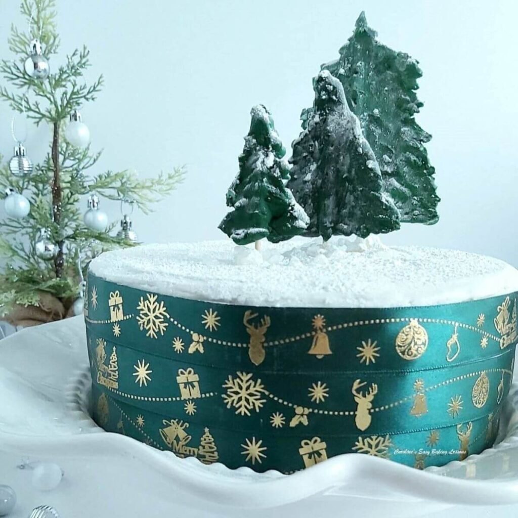 White royal iced deep Christmas cake with bottle green ribbon on sides, homemade trees and tree to the back in a white setting - cropped..