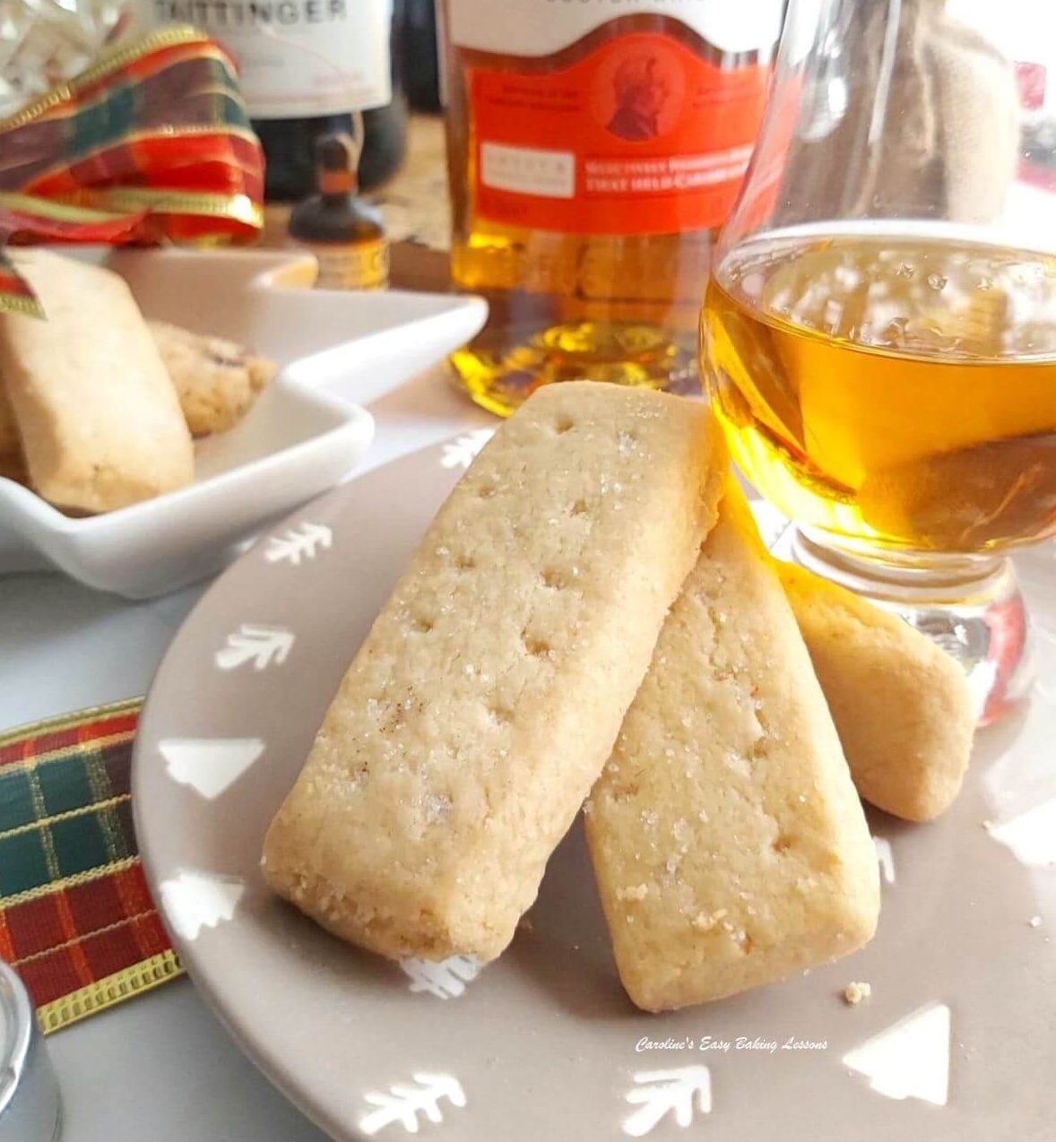 Close photo fo 2 sticks of Scottish shortbread cookies on a Xmas plate with whisky glass and bottle and party food behind.