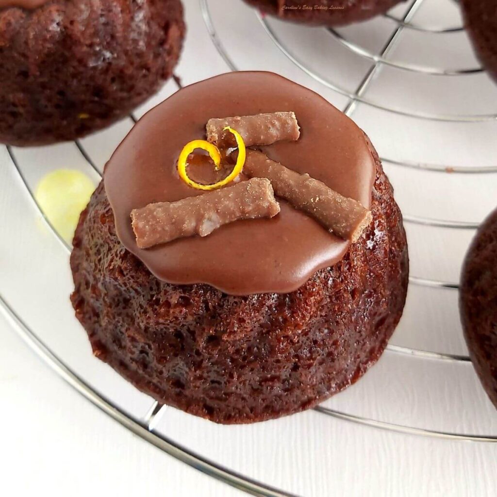 Extra close above shot of a mini chocolate bundt cake on a cooling rack, with chooclate glaze on top, orange zest twirl and broken matchmaker chocolate pieces.