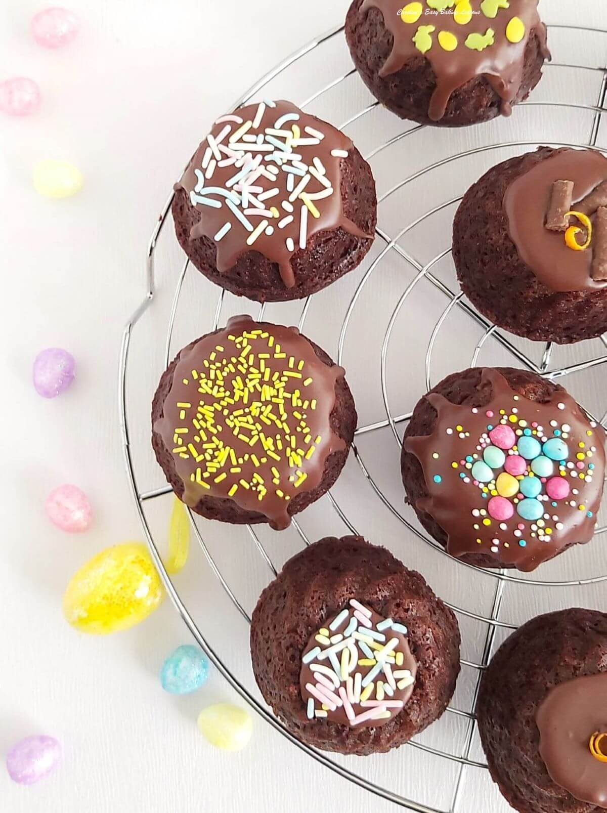 Partial shot, overhead of a white table with cake cooling rack and 7 mini chocolate bundt cakes with glaze and sprinkles and mini eggs to the sides.