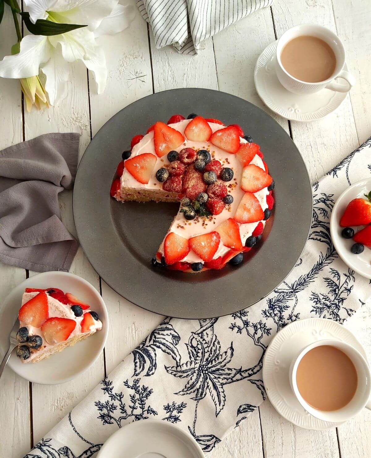 Overhead shot of white dinner table with linen and crockery surrounding a stawberry, blueberry and raspberry covered cream layer cake called Summer Berry Griestorte.