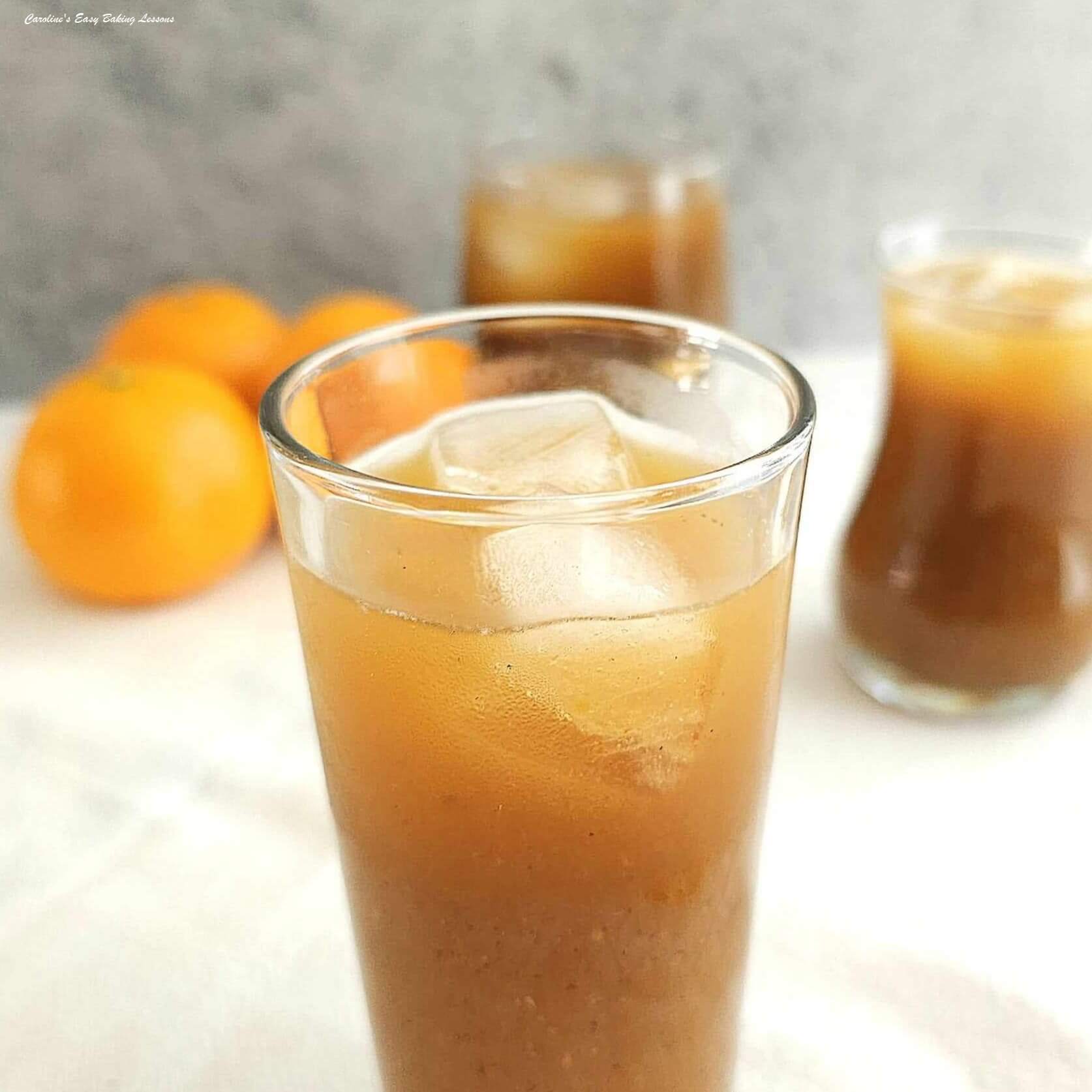 Close shot of a tall slim glass with persperation, and filled with brown/orange coloured fruit ice tea with ice cubes, and oranges and glassed to the background.