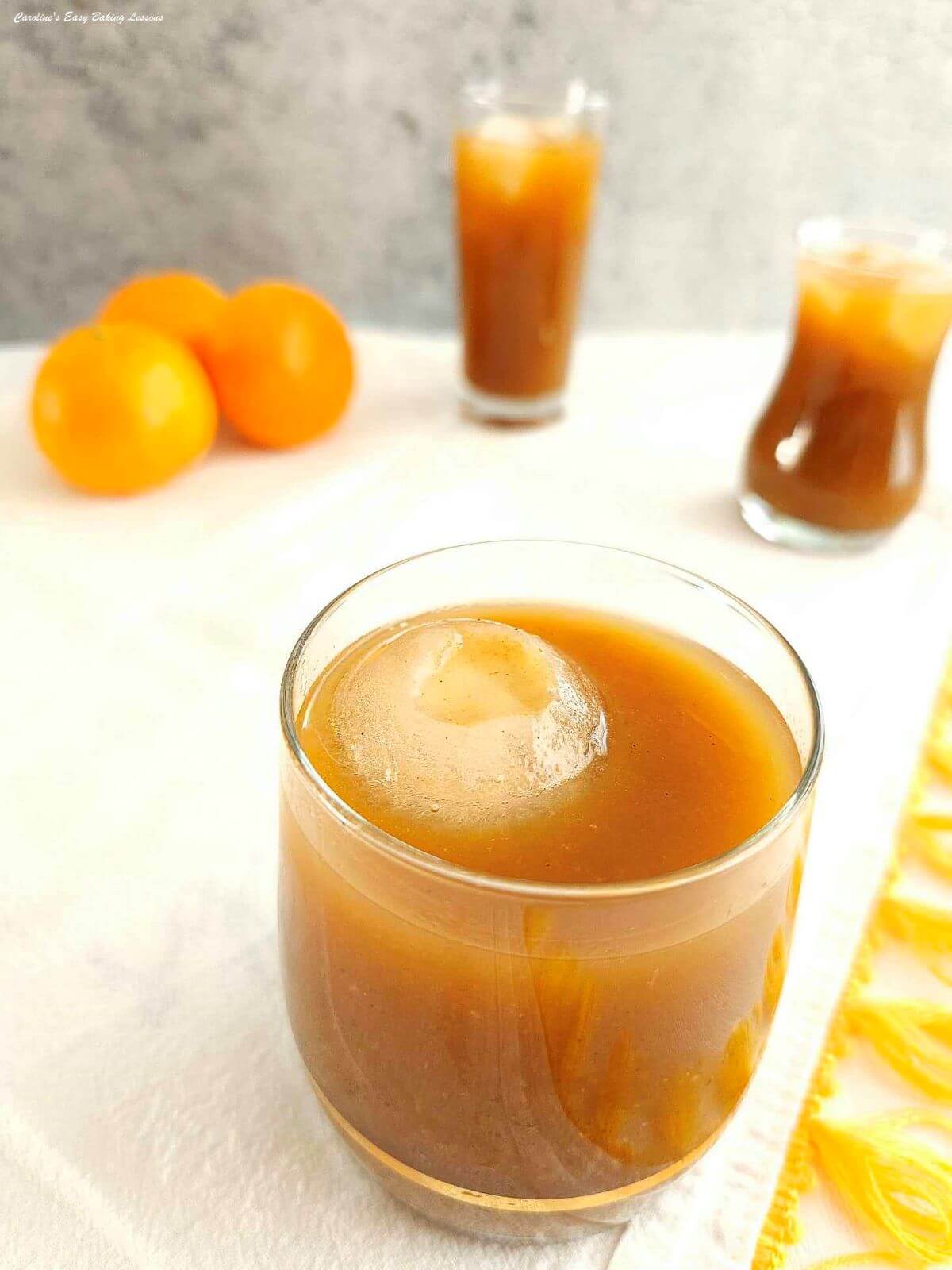 Whisky glass with orange/brown citrus tea and extra large round ice cube, with oranges and 2 different sized glasses to the background.
