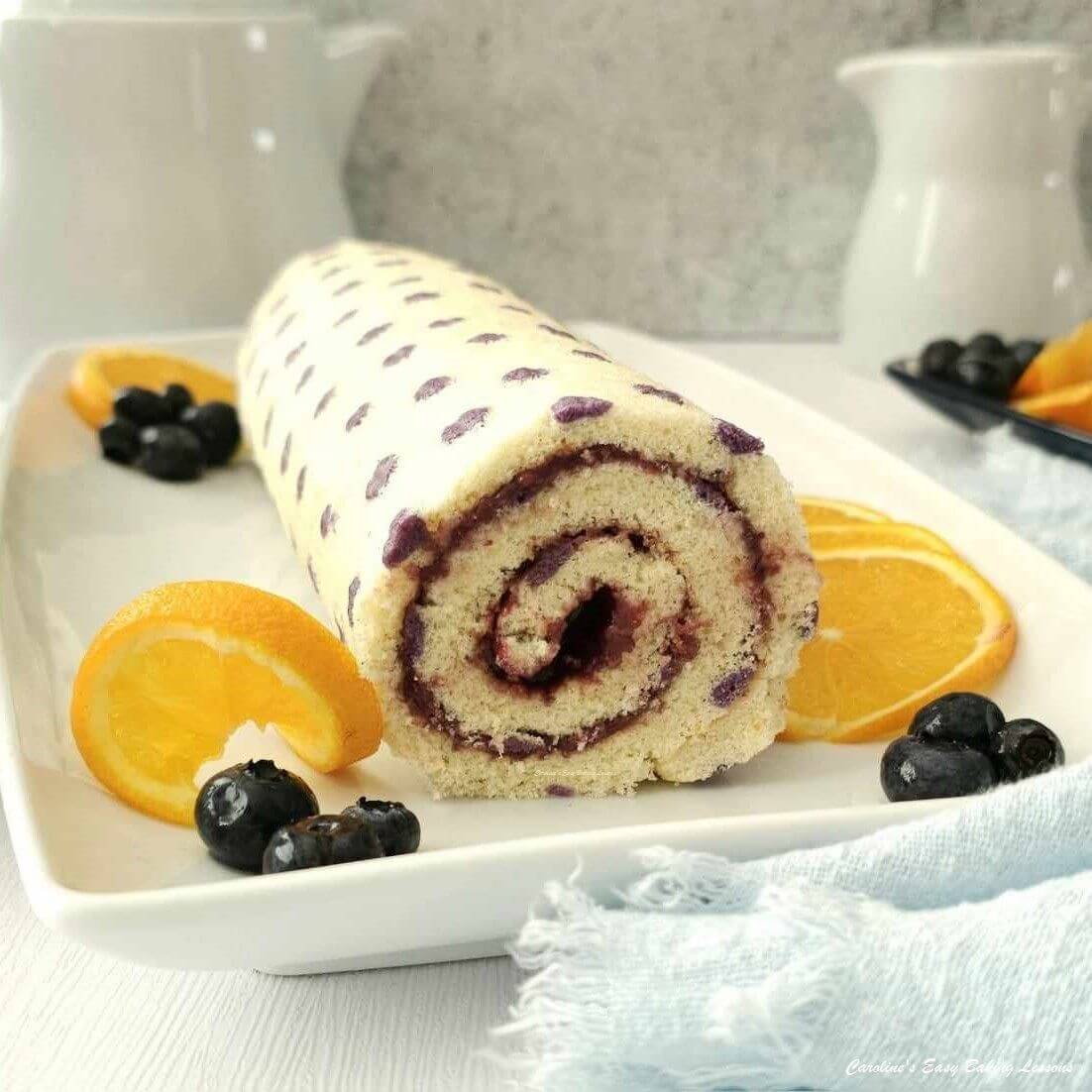 45 degree Shot of end of a purple dot patterned Swiss roll/jelly roll cake with bluberries and orange garnish.