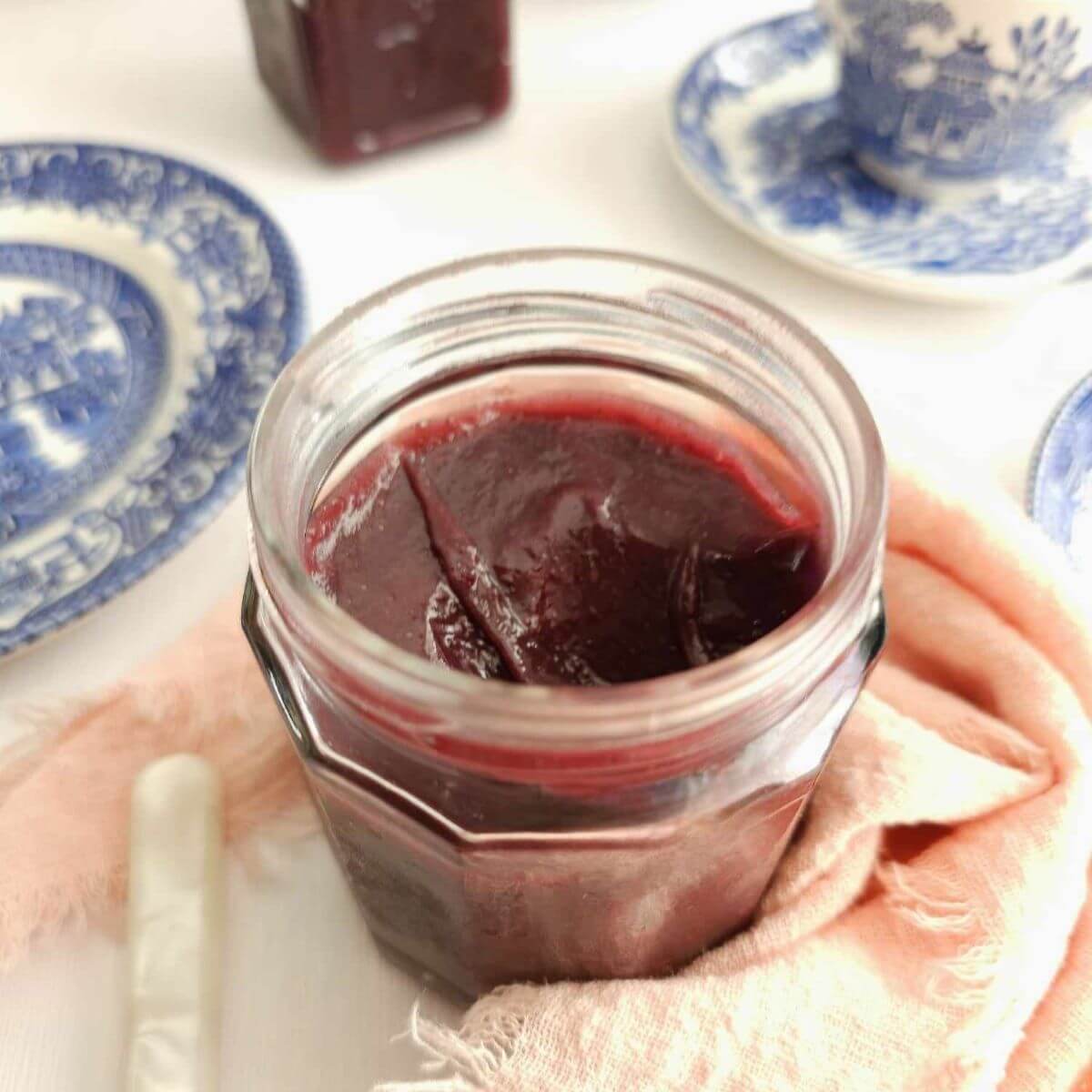 Close phhoto of open jar of purple coloured bluberry orange curd, with plates surrounding it.