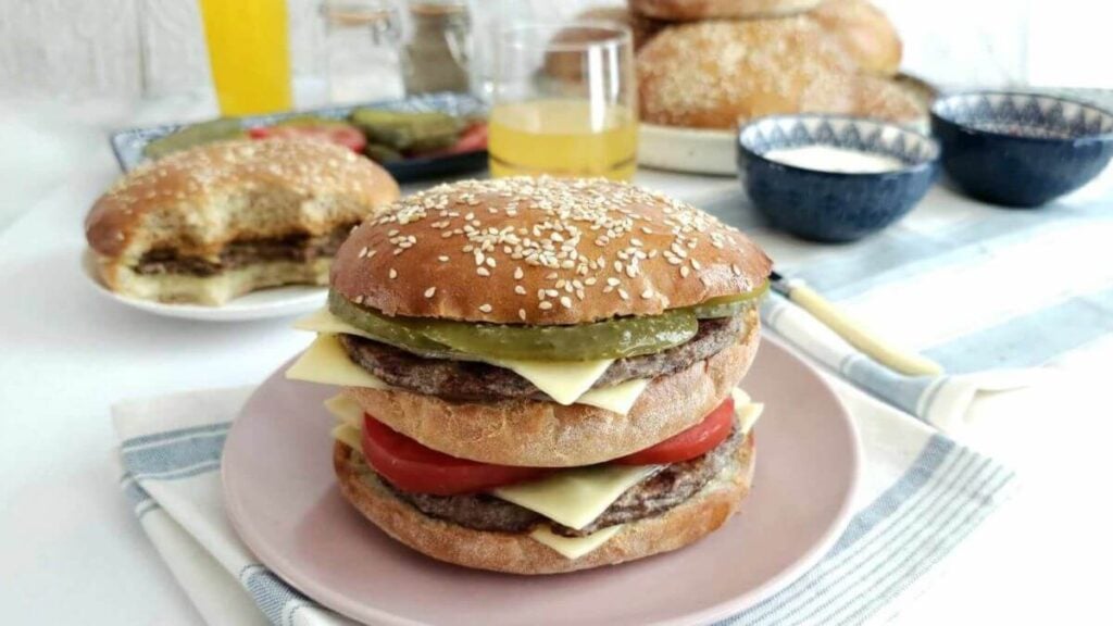 Shot of tall homemade double decker cheese burger with deep golden brown seeded top, cheese, tomoato and gerkins visible, on a pale pink plate with burger and drnks to the background.