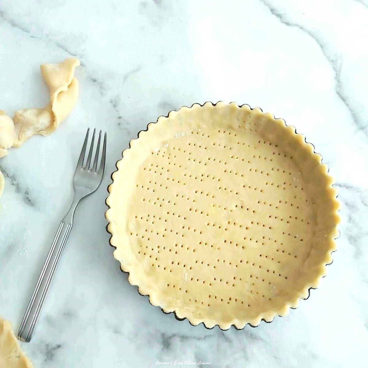 Overhead shot of a round pie tin lined with pastry, excess pastry& fork to the side, and tiny holes in the base of the uncooked pastry case called 'docking'.