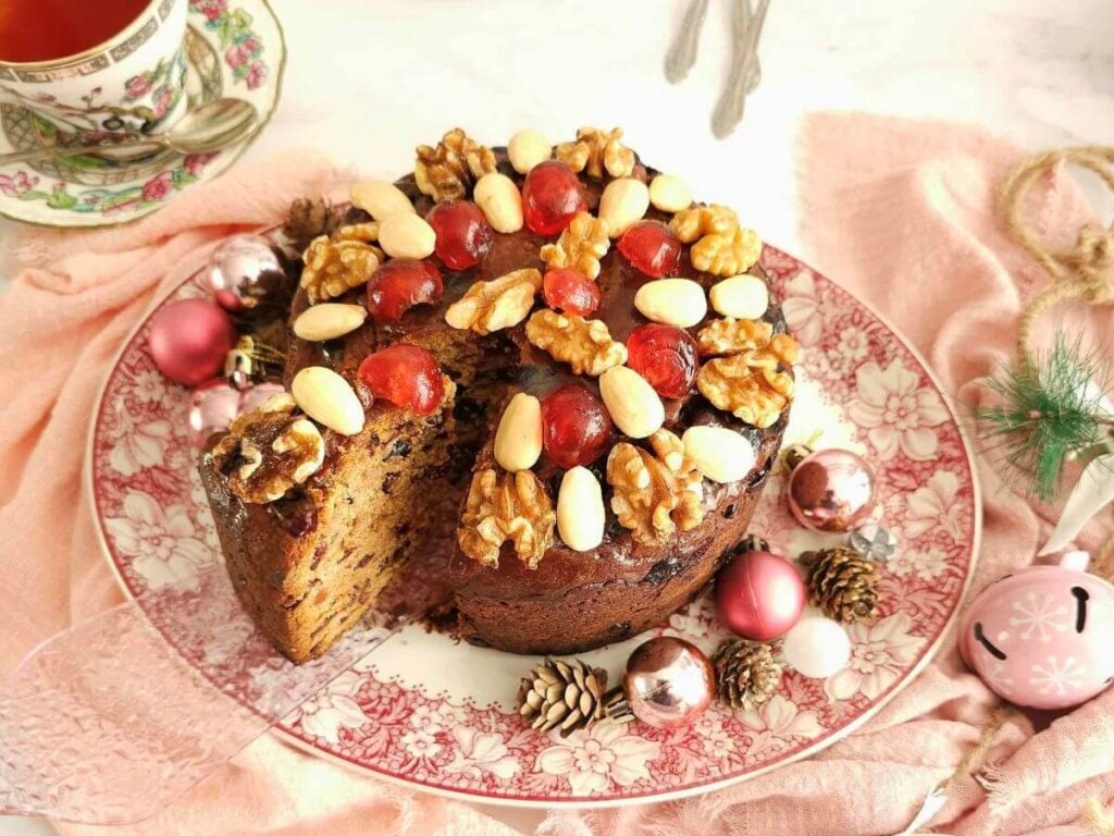 Overhead shot of a nut and cherry topped fruit cake, on table with pink napkin, crockery and pink Christmas decorations and a slice about to be removed.