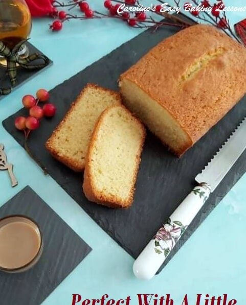 madeira cake with 2 slices cut on black slate board on pale blue background with drinks and red berries.