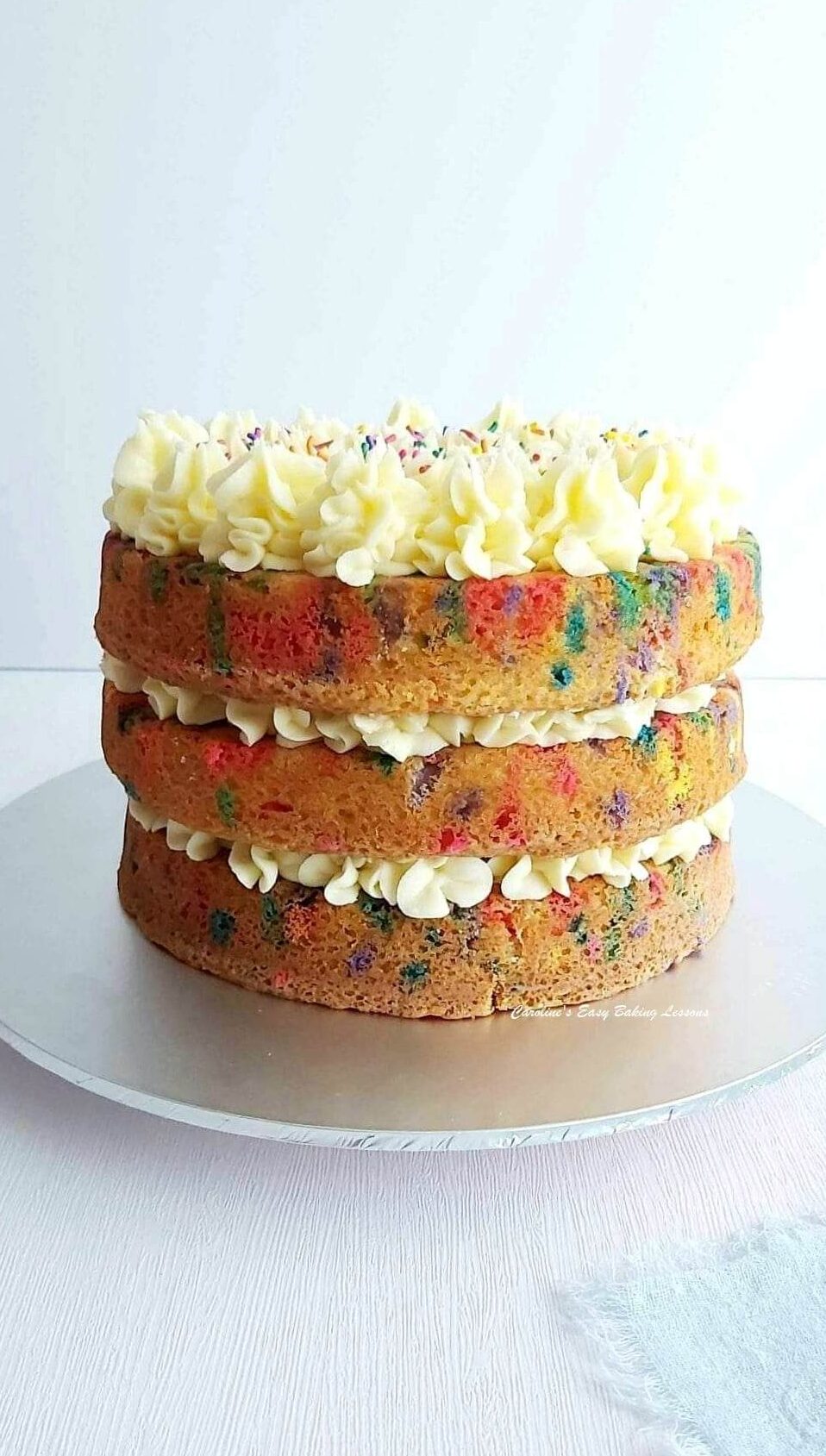 Front on long shot of a 3 layer funfetti sprinkle cake with piped buttercream on a white table and background.