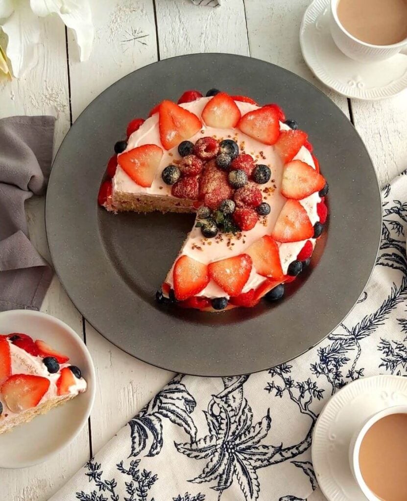 Overhead shot of white dinner table with linen and crockery surrounding a stawberry, blueberry and raspberry covered cream layer cake called Summer Berry Griestorte.