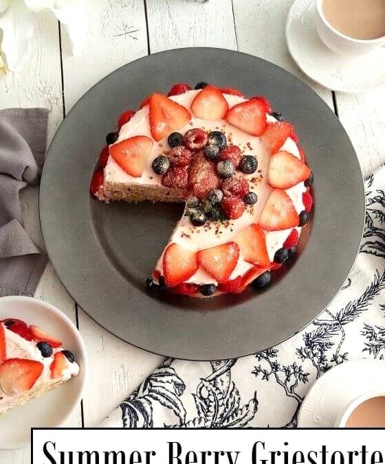 Overhead shot of white table with blue white napkins, white flowers, tea cups with tea and large strawberry, raspberry & blueberry covered cream layer cake in centre and text Summer Berry Griestorte.