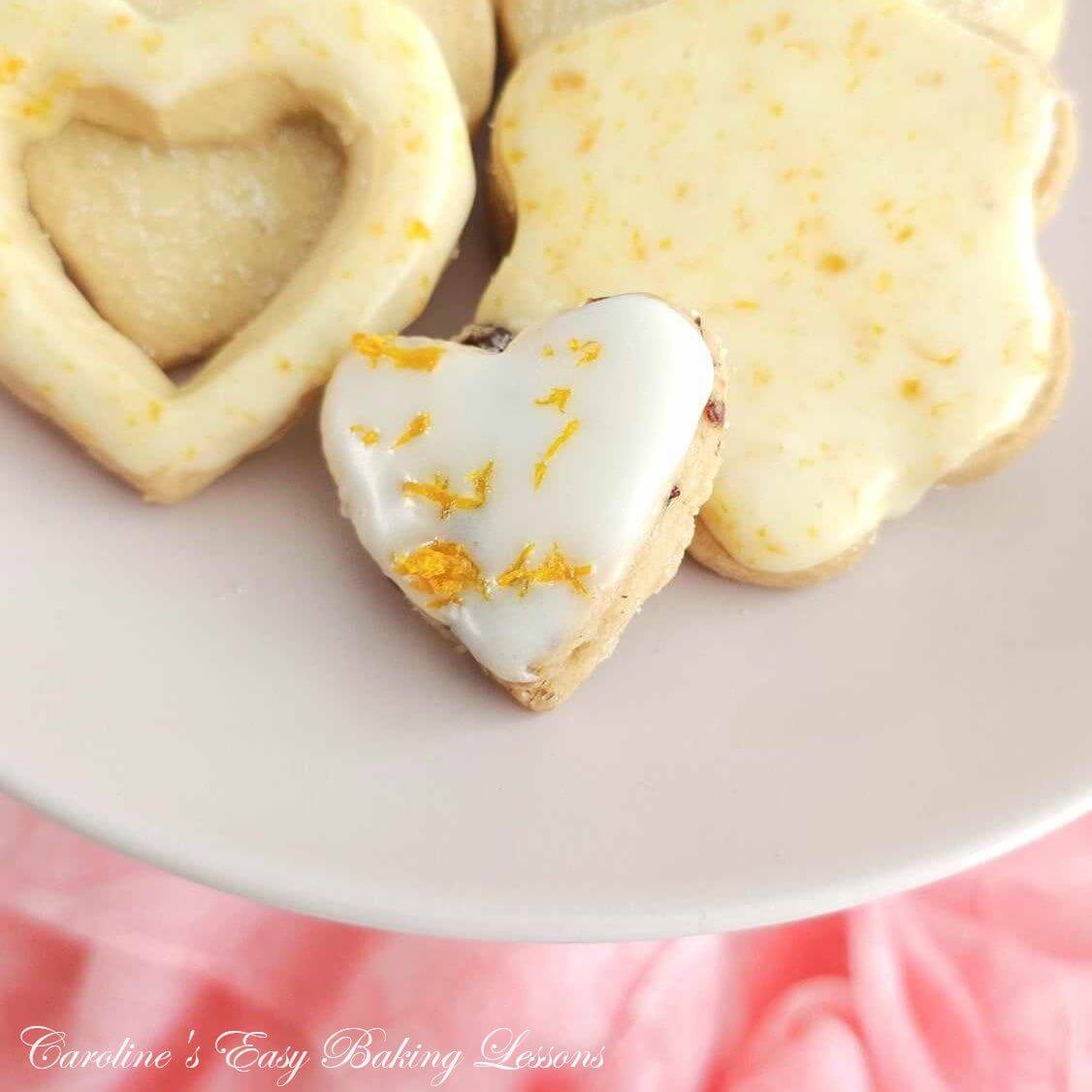 Extra close partial shot of pink plate and some heart & flower shaped Scottish shortbread, one with cut out heart centre and orange zest glaze, around a pink napkin.