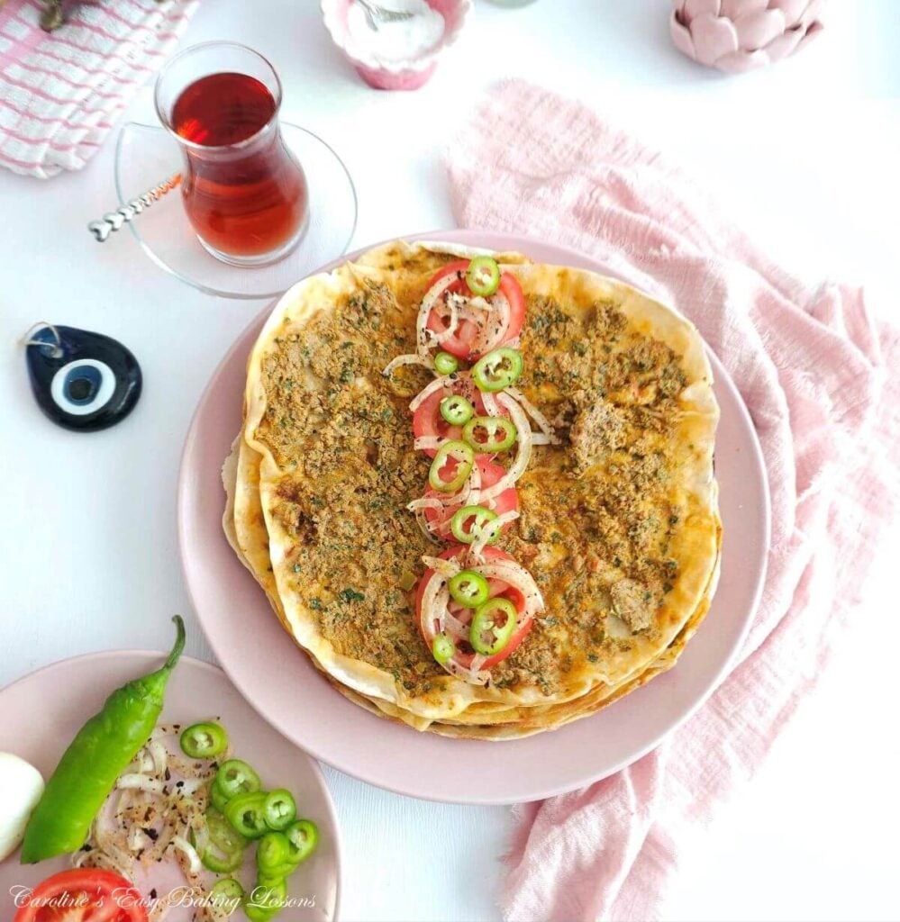 Overhead shot of white table with pink crockery and napkin, Turkish tea cup, and in centre a ground meat topped Lahmacun - flatbread with slices of tomoato, onion & pepper on top.
