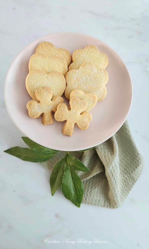 Overhead photo of a pink plate on a white marble top, with shaped Scottish shortbread biscuits, and 2 in clover shape, with green napkin and follage leaved.
