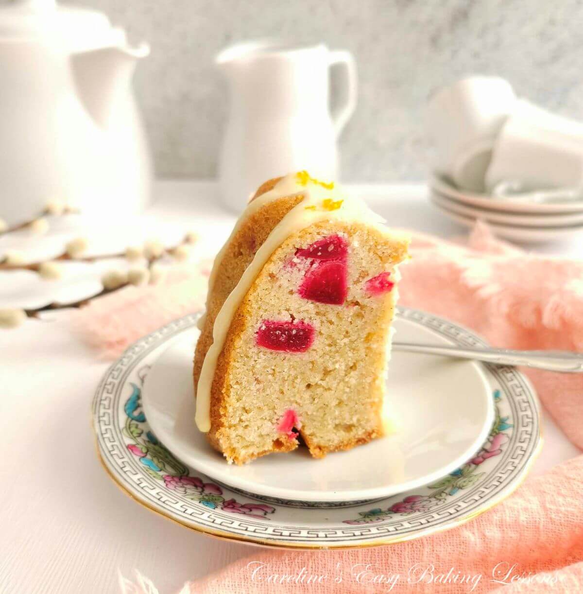 Close front shot of a tall slice of red plum filled bundt cake with glaze drips, served on aplate with fork and pink napkin to the side, white crockery to the back.