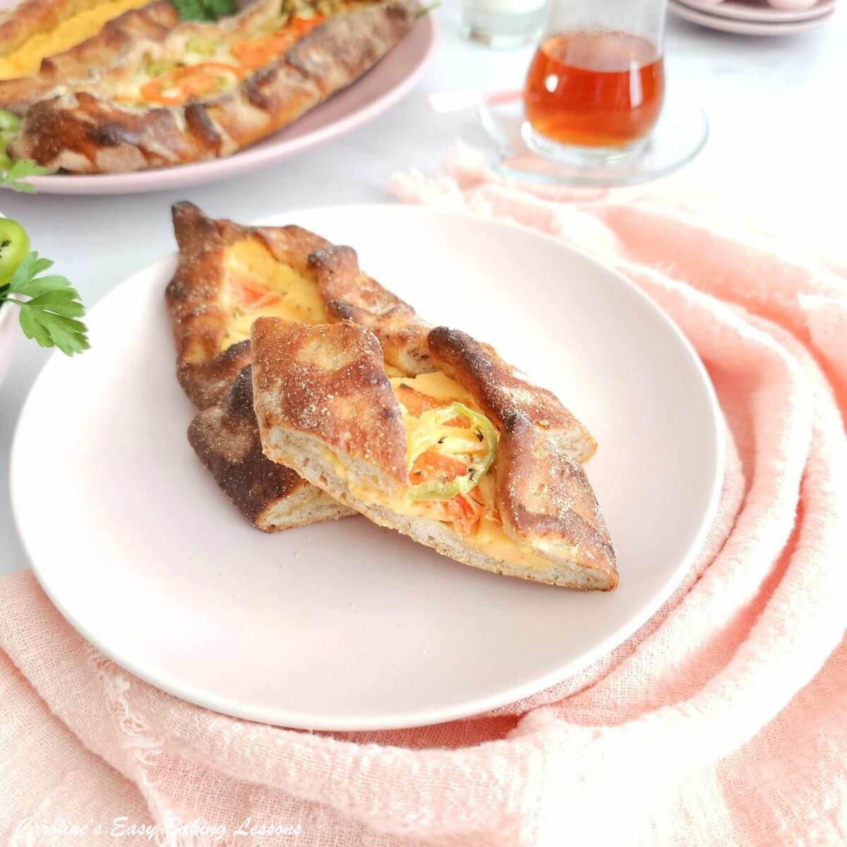Photo of several slices of a cheese and tomato topped Turkish Pide pizza, ovan long shaped, on a pink plate, with pink napkin and Turkish tea glass to the background.