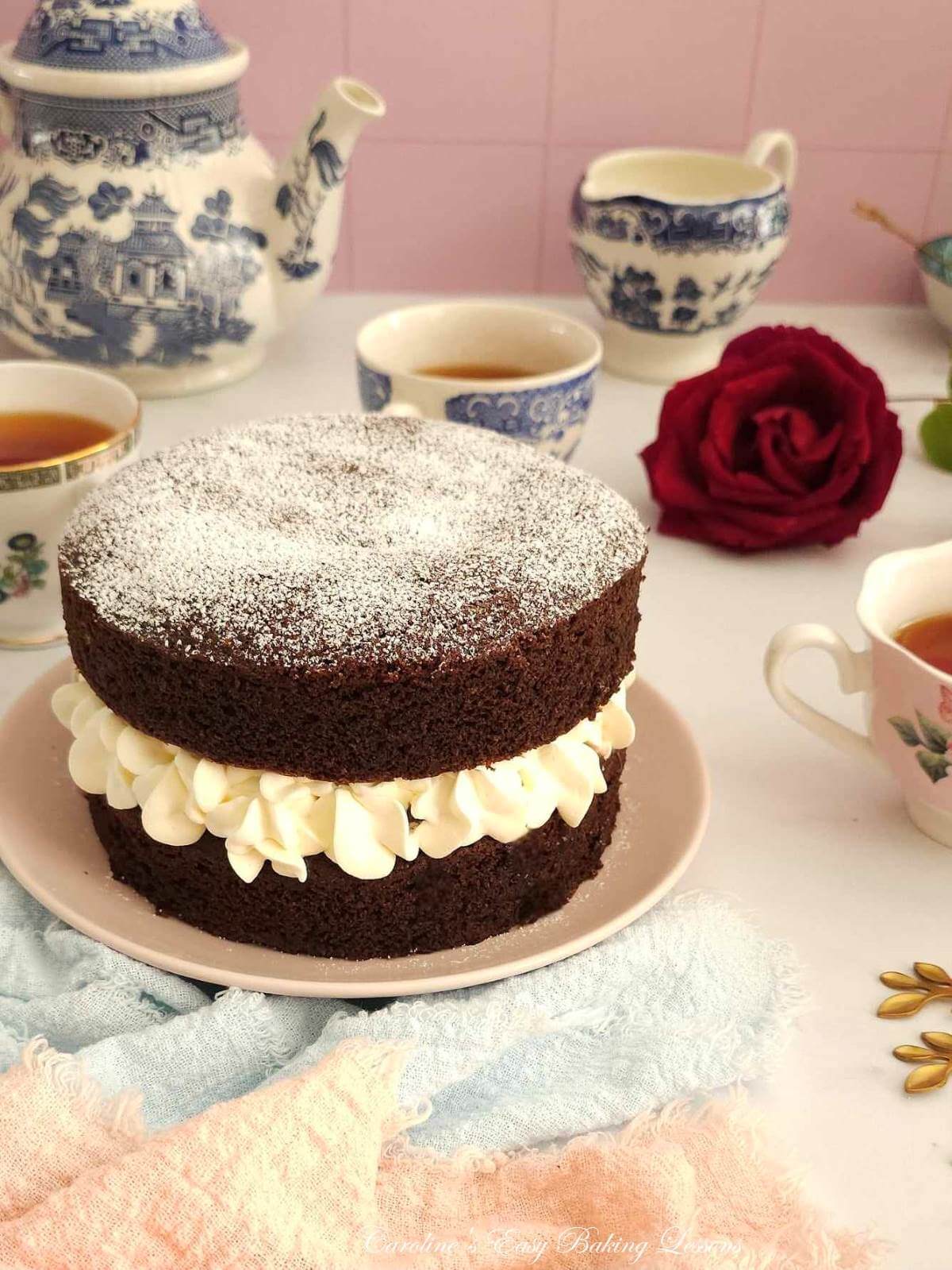 Angled shot of a chocolate Victoria sandwich cake on pink plate, with blue, pink and white crockery and teapot to the background.
