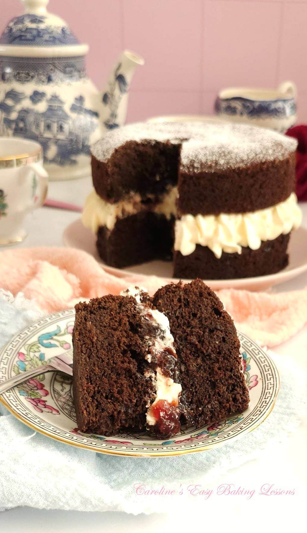 Close photo of slice of chocolate Victoria sandwich cake with piped cream and cherry jam filling, and the whole cake to the background.