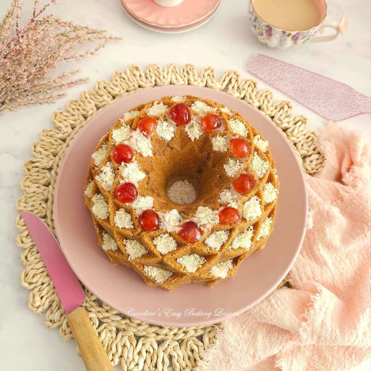 Overhead shot of pink crockery and napkin, with fancy bundt cake, with glaze, coconut and cherry topped bundt cake.