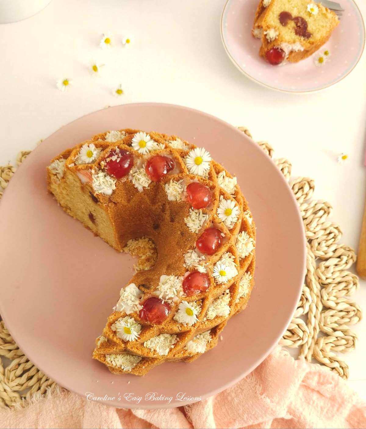 Overhead shot of half a fancy bundt cake, Bakewell Bundt), with glaze, coconut, cherries and daisy flowers, on a white tabletop with napkin & slice to the background.