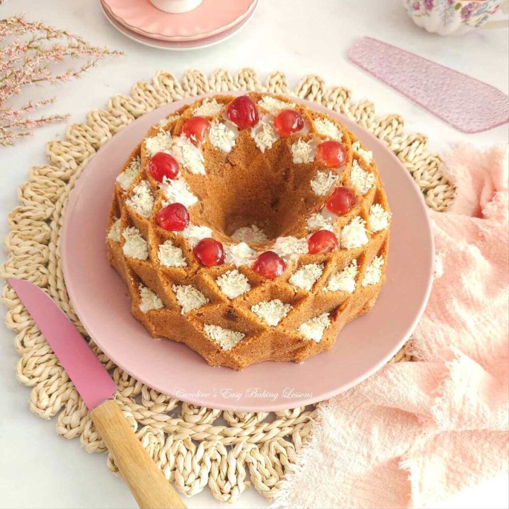 Overhead shot on a white table, with pink crockery and napkin, & a Bakewell Bundt cake in the centre, decorated with cherries, glaze, & coconut.