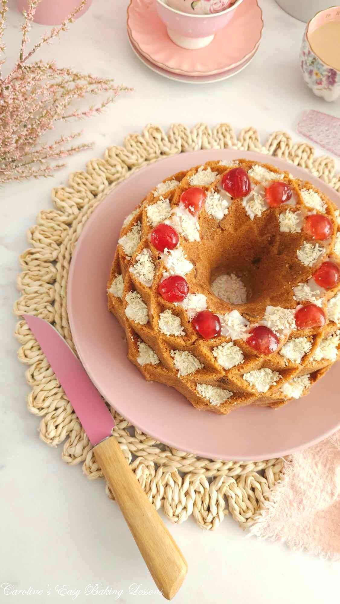 Overhead shot of a white table with pink crockery & line, with a partial view of a fancy bundt cake topped with coconut and cherries.