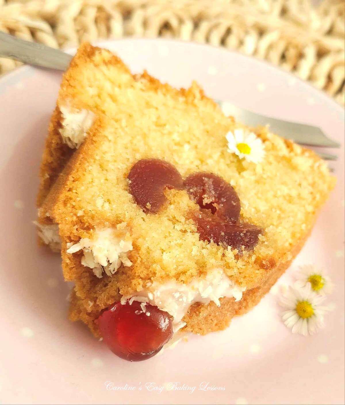 Extra close shot of a slic eof Bakewell Bundt cake, filled with cherries, topped with a cherry, glaze & coconut, served on a pink plate.