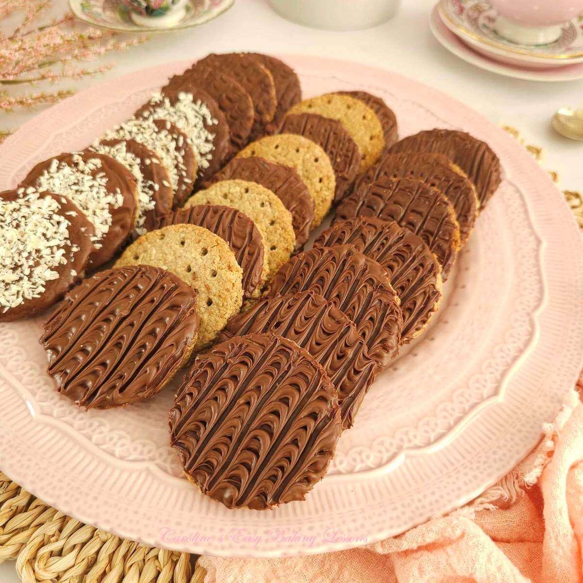 Photo of white tble with pink crockery and linen, with large plate of 3 rows of homemade chocolate, plain & coconut topped digestive biscuits.