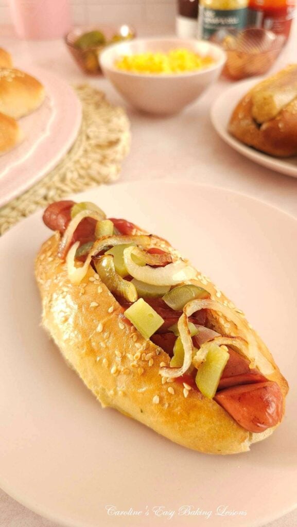 Pink coloured table with pink plate of home made hot dog bun, filled with hot dog, and fillings, more buns to the background.