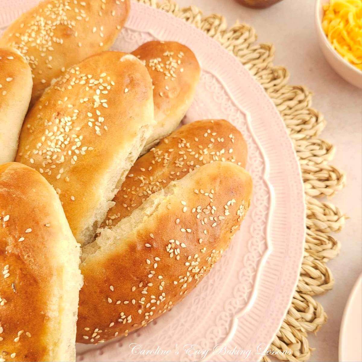 Partial shot of a pink plate of golden homemade sesame topped hot dog buns, on a pink plate.