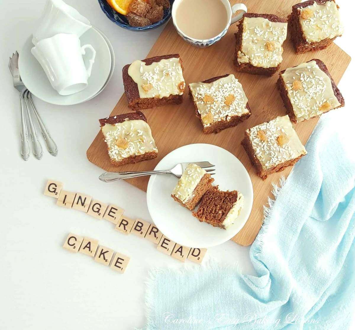 Overhead photo on white table with blue napkin and crockery, and a wooden chopping board with glazed cake squares, and scrabble letters spelling out 'gingerbread cake'.