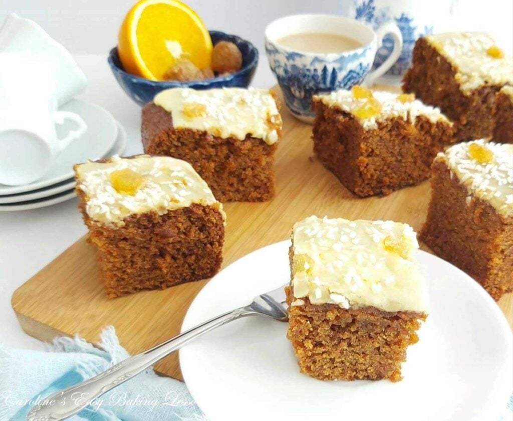 Front angled photo of some gingerbread cake square son chopping board and one served on white plate at front.