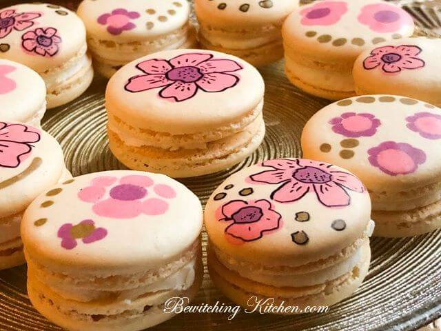 Photo of plate of several pink macarons, with painted flowers on top, and text 'Bewitching Kitchen.com'.