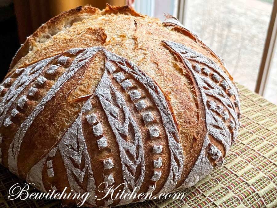 Close front on shot of a well detailed scored patterned sourdough round bread, with text 'bewiitching kitchen.com'.
